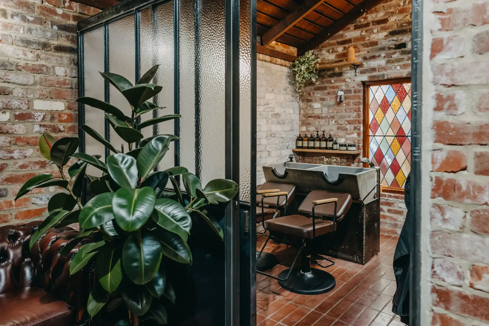 Inside George & Ivy Fremantle: heritage red brick wash bay with stained glass window, leather Chesterfield sofa and a rubber plant by the basins