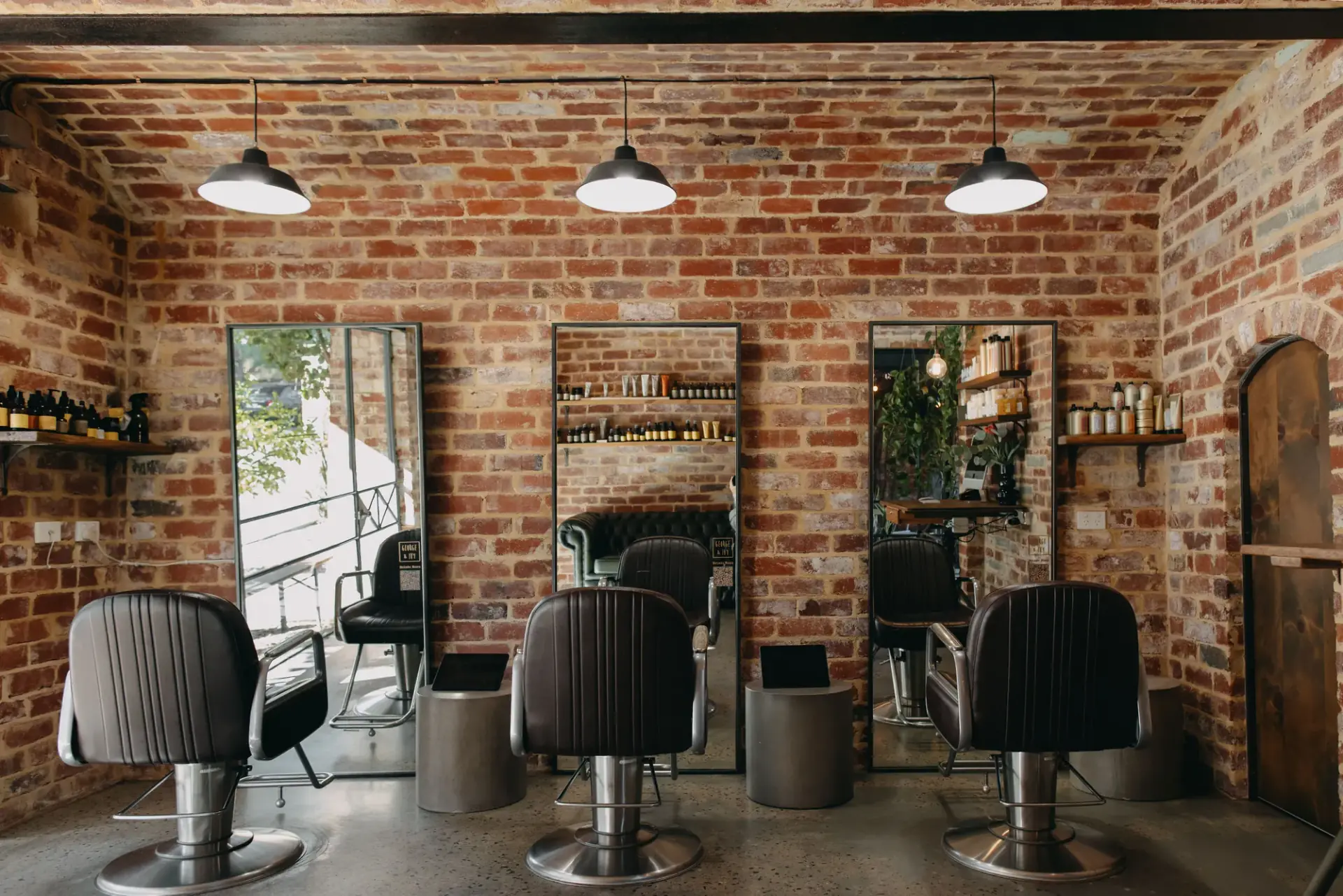 Inside George & Ivy Fremantle: three leather styling chairs against an exposed red brick wall with industrial pendant lights and floor‑to‑ceiling mirrors