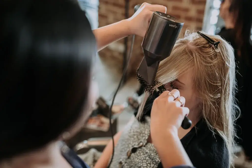 A stylist blow-drying a blonde client’s hair with a round brush at George & Ivy, in front of the salon’s exposed red brick wall