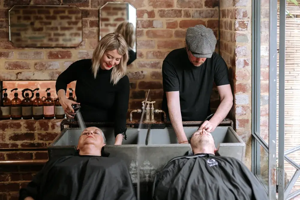 Two George & Ivy stylists washing clients side by side at the salon’s vintage galvanised trough basin, with a shelf of amber Oway bottles on the brick wall behind