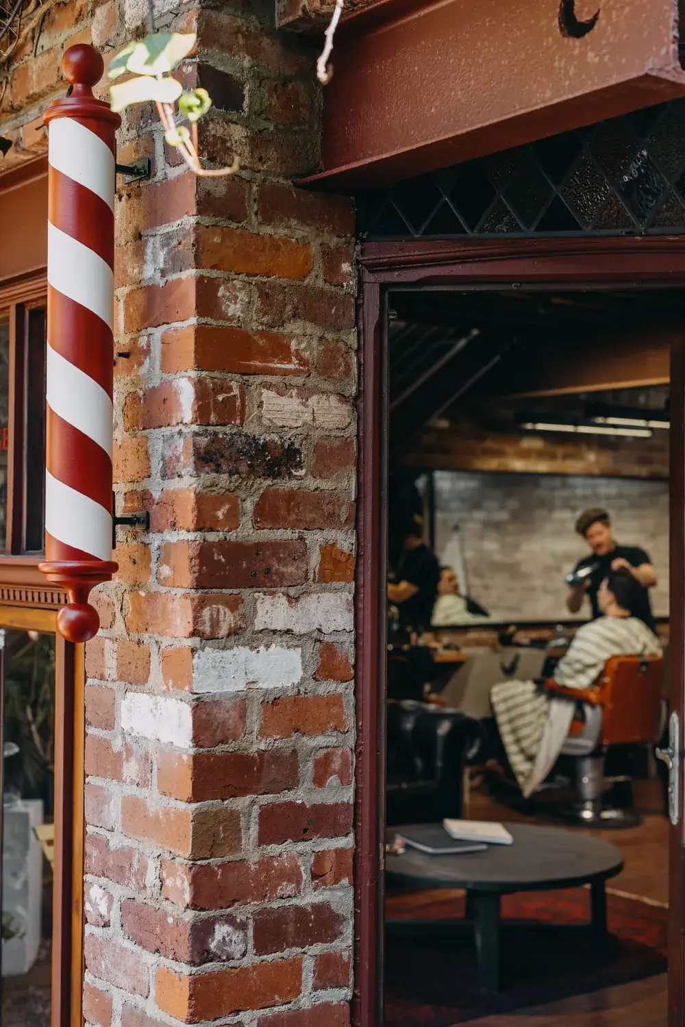 The Two Georges barbershop in Fremantle: a red and white barber pole mounted on the red brick shopfront, with a barber styling a client in the chair inside